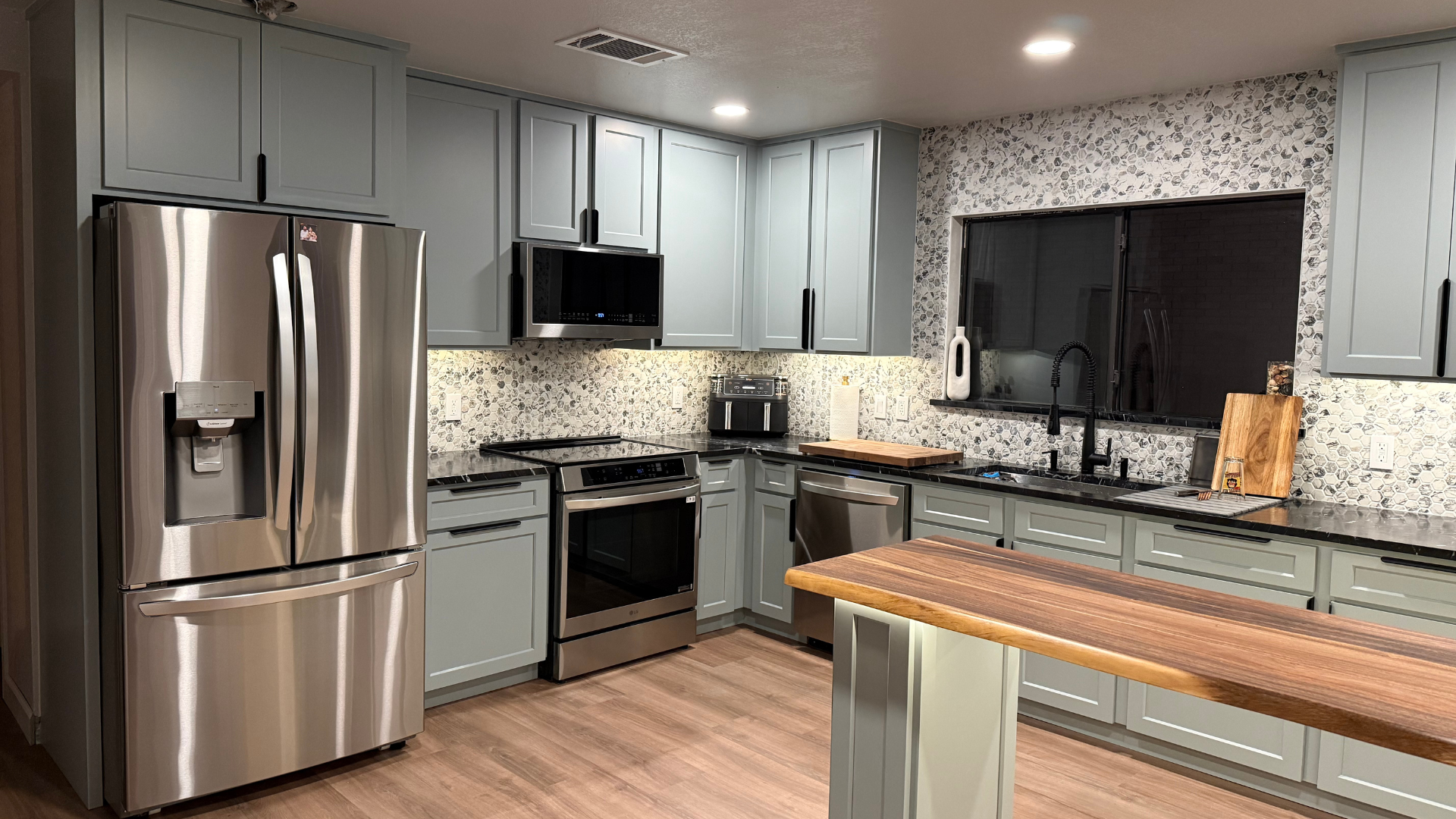 Refinished sage-green kitchen cabinets with stainless appliances and marble-pattern backsplash